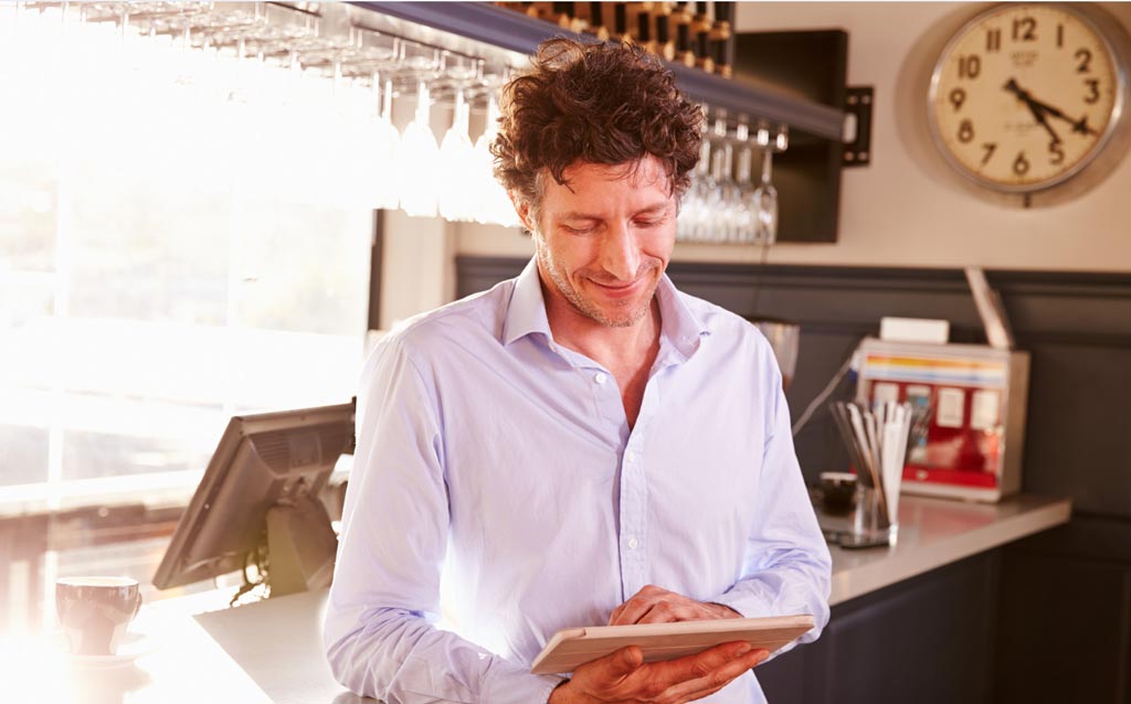 Restaurant owner leaning against a counter and looking at his tablet and smiling